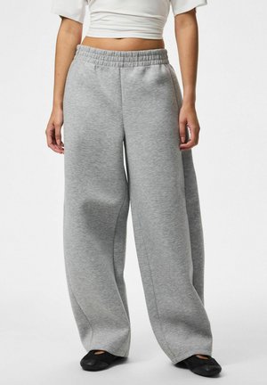 Vrouw draagt lichtgrijze wijde joggingbroek, wit cropped topje en zwarte instapschoenen, staand tegen een witte achtergrond.