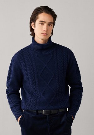 Jonge man met donker haar die een marineblauwe kabelgebreide coltrui en een donkere broek draagt, met handen in de zakken tegen een grijze achtergrond.