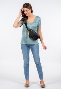 Borsa a tracolla nera in tessuto con superficie testurizzata, con zip gialla, indossata a tracolla da una donna in una blusa azzurrino chiaro e jeans blu.
