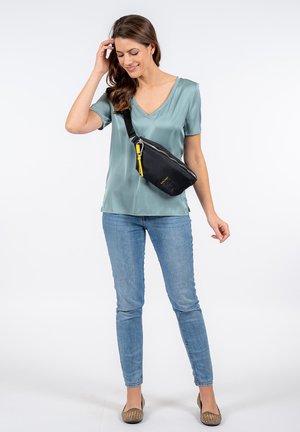 Sac en toile noire avec surface texturée, zippe en jaune, porté en bandoulière par une femme en blouse bleu clair et jeans bleus.