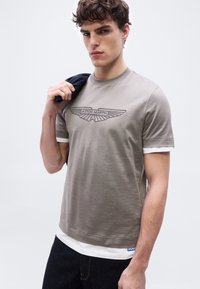 Νεαρός άνδρας με σγουρά μαλλιά φοράει γκρι μπλουζάκι Aston Martin πάνω από λευκό μακρυμάνικο, κρατώντας σκούρο σακάκι πάνω στον ώμο του μπροστά σε απλό φόντο.