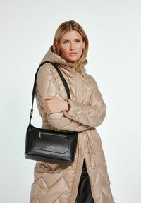 Czarna skórzana torba na ramię z zamkiem błyskawicznym, prostokątny kształt, gładka faktura i metalowe akcenty, noszona z beżową kurtką puchową.