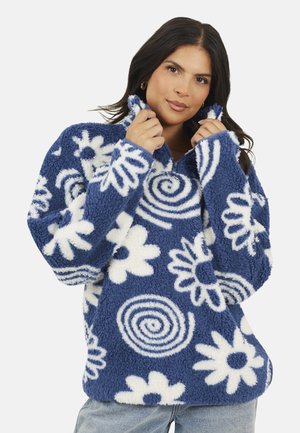 Frau, die den Kragen eines blauen Fleece-Pullovers mit weißen Spiralen und Blumenmustern anpasst, trägt helle blaue Jeans.