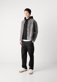 Grå denimjakke med lommer, svart hettegenser under, mørke bukser og hvite sko med beige detaljer. Modellen står i et nøytralt rom.