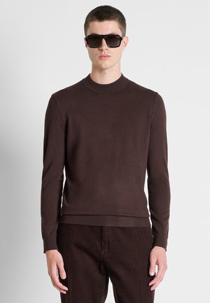 Junger Mann trägt einen dunkelbraunen Pullover und Cordhosen mit großen schwarzen Sonnenbrillen, er steht vor einem schlichten hellen Hintergrund.