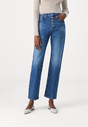 Person trägt hoch taillierte blaue Jeans mit sichtbaren vorderen Knöpfen, beige geripptes Langarmshirt und weiße spitze flache Schuhe, stehend auf weißem Boden.