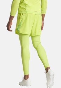 Neon gele sportoutfit bestaande uit een strakke lange mouw top, losse shorts en bijpassende leggings. Witte voetbalschoenen zijn zichtbaar.