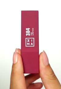 Un tube de rouge à lèvres bordeaux étiqueté "384 LE ROUGE À LÈVRES" avec un texte blanc. Texture lisse, forme rectangulaire, tenu entre les doigts.