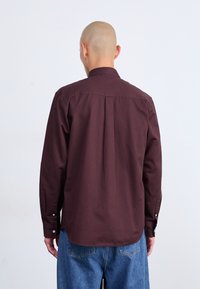 Camisa burdeos de manga larga con cuello abotonado y detalles en la yoke, combinada con pantalones de mezclilla azul claro. Textura de algodón suave.