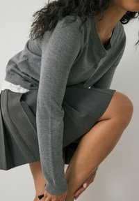 Grauer Strickcardigan mit Knöpfen, über ein weißes Hemd getragen, kombiniert mit einem gefalteten dunkelgrauen Rock mit glatter Textur.