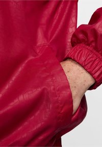 Chaqueta ligera roja con tejido texturizado. Dispone de puños elásticos y un bolsillo lateral, con detalles sutiles de patrón en toda la superficie.