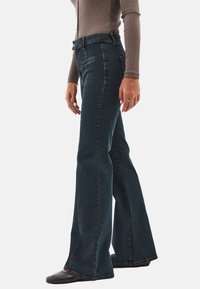 Jeans acampanados de color azul oscuro con cinco bolsillos, costuras doradas, lazos para cinturón y una cintura alta. La tela tiene un acabado suave y ligeramente texturizado.