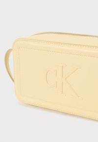 Sárga, téglalap alakú bőr táska, dombornyomott "CK" logóval, cipzáras záródással és varrási részletekkel. Kisebb oldalsó fogantyúval rendelkezik.