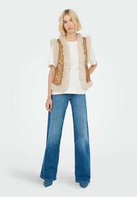 Braune Weste mit flauschigem beigem Besatz und gestickten Mustern, getragen über einem weißen Hemd und kombiniert mit weit geschnittenen blauen Jeans.