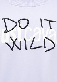 Ανοιχτό μωβ ύφασμα με την επιγραφή "DO IT WILD" σε έντονα μαύρα γράμματα πάνω από αχνό λευκό κείμενο.