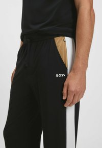 Close-up van een persoon die zwarte trainingsbroek draagt met witte en tan zijkanten, een "BOSS" logo en een zwarte trekkoord taille.