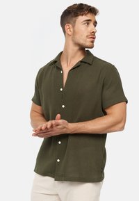 Camicia corta verde oliva, tessuto testurizzato, con bottone davanti, colletto piatto, bottoni bianchi e un piccolo logo etichetta sull'orlo.