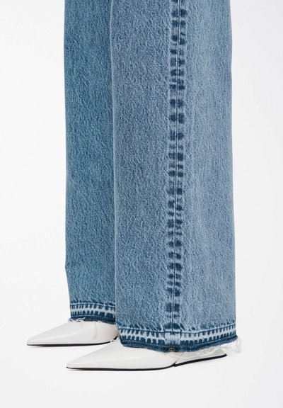 Los pantalones de mezclilla azul de pierna ancha presentan dobladillos deshilachados y una textura desgastada, combinados con tacones blancos puntiagudos para crear un contraste.