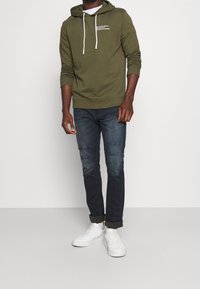 Olivgrüner Hoodie mit weißen Kordeln und kleingedrucktem Text. Kombiniert mit dunkelblauen Jeans und weißen Sneakers. Einfacher, lässiger Look.