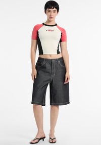 Cropped shirt in white, red, and green, paired with dark denim bermuda shorts with contrasting white stitching. Model wears black sandals.