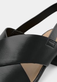 Κοντινή λήψη μαύρου δερμάτινου σανδαλιού ανοιχτής μύτης με σταυρωτές λουρίδες και μπεζ εσωτερική σόλα σε λευκό φόντο.