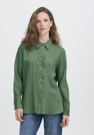 Giovane donna con capelli castani ondulati, che indossa una camicia verde a maniche lunghe con bottoni e jeans blu, in piedi contro uno sfondo bianco semplice.