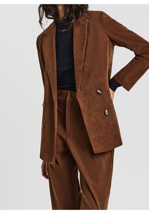 Femme portant un blazer en velours côtelé marron à double boutonnage et un pantalon assorti avec un col roulé côtelé noir, posant avec une main sur la hanche.
