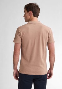 Béžové polo tričko s krátkými rukávy, strukturovaná tkanina a límeček. Má malé logo na levém rukávu. Model je otočen zády.