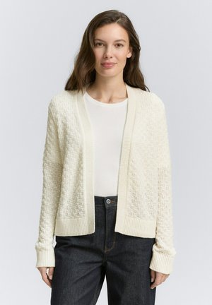 Cardigan en tricot crème avec un motif texturé, ouvert sur le devant, avec des poignets et un ourlet côtelés. Porté par-dessus une chemise claire et un jean noir.