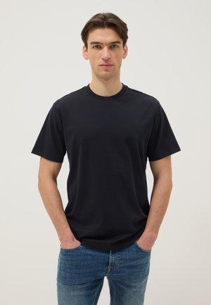 Giovane uomo in piedi con le mani in tasca che indossa una maglietta nera a maniche corte semplici e jeans blu su uno sfondo bianco.