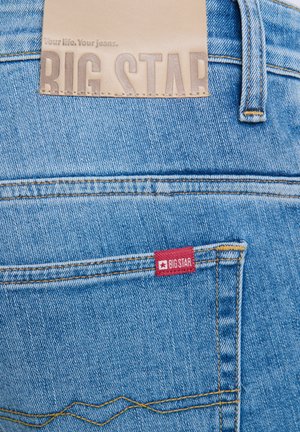 Svetle modré džíny vyrobené z denimu, s béžovou koženou nášivkou s nápisem "BIG STAR" a červeným štítkem se stejným logem na zadní kapse.