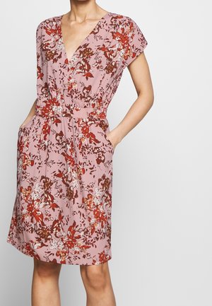 Femme portant une robe rose sans manches avec imprimé floral rouge et blanc, col en V, taille élastique et mains dans les poches latérales sur fond uni.