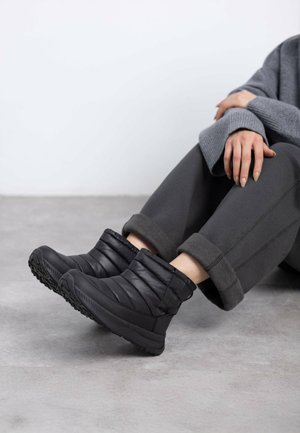 Botines negros aislados hasta el tobillo con textura acolchada, suela de goma y una lengüeta en la parte posterior. Se pueden ver unos pantalones grises con puños y un suave suéter gris.