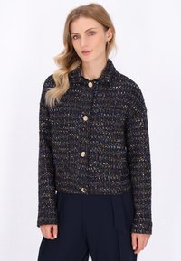 Una donna bionda posa indossando un maglione scuro texture con accenti metallici, dotato di colletto e bottoni dorati, su uno sfondo bianco semplice.