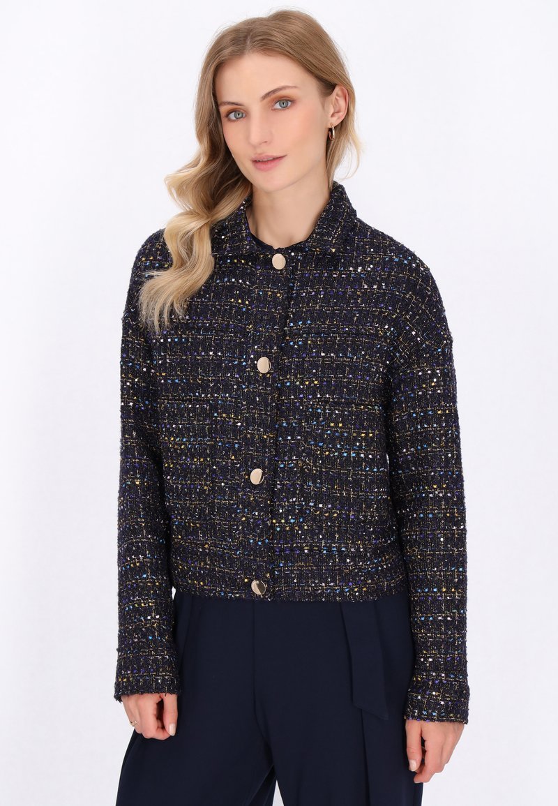 Una donna bionda posa indossando un maglione scuro texture con accenti metallici, dotato di colletto e bottoni dorati, su uno sfondo bianco semplice.