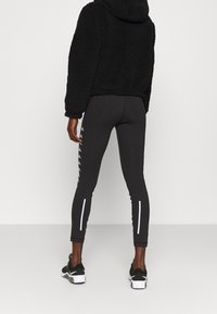 Felpa nera pelosa con cappuccio, abbinata a leggings neri aderenti con strisce bianche sui lati, indossata con scarpe da ginnastica nere.
