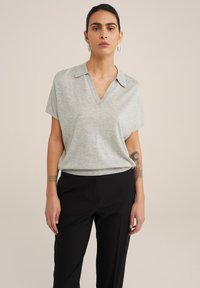 Šedý polo top s krátkým rukávem, má v�ýstřih do V a límec, jemná textura, spárovaný s černými kalhotami. Viditelné tetování na pažích dodává detail.