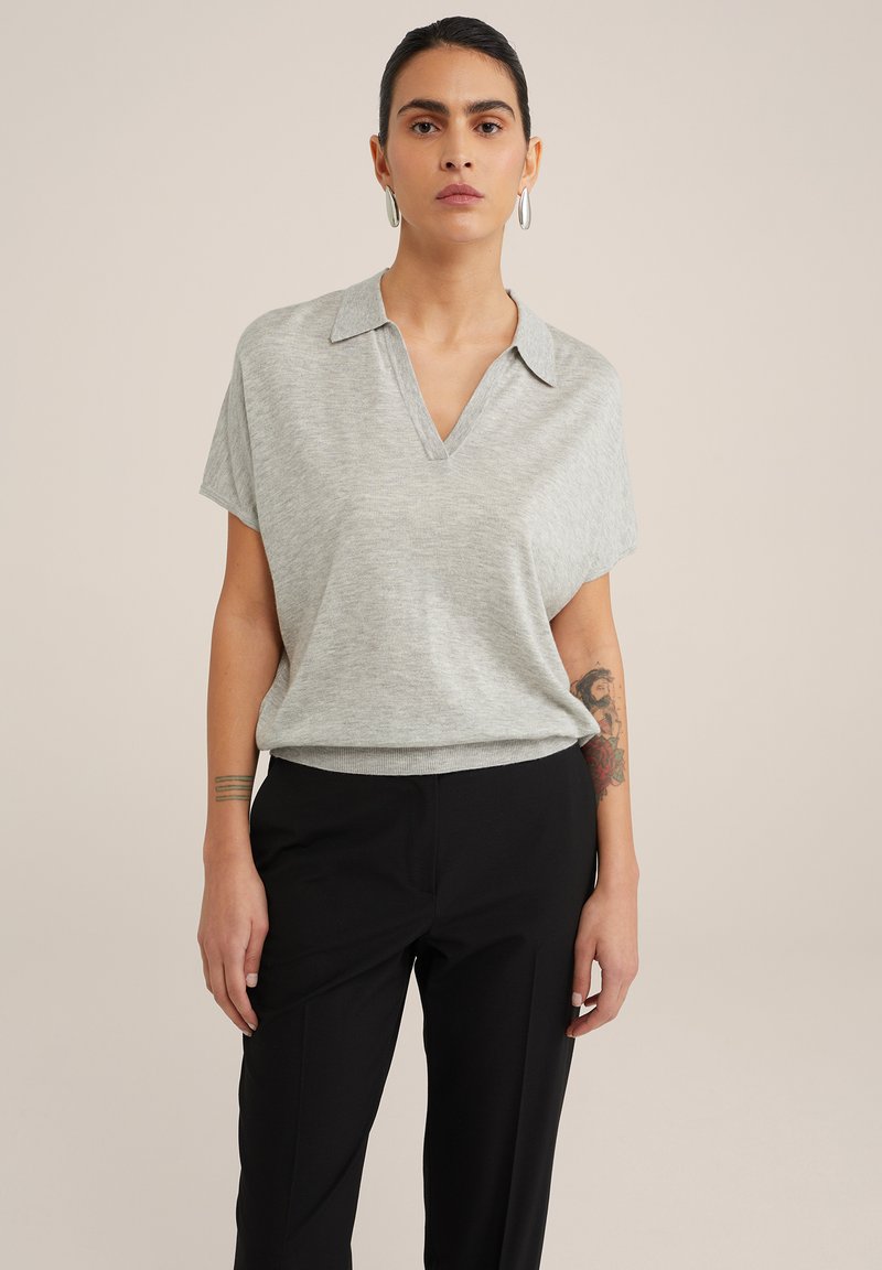 Šedý polo top s krátkým rukávem, má výstřih do V a límec, jemná textura, spárovaný s černými kalhotami. Viditelné tetování na pažích dodává detail.