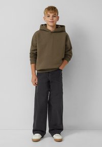 Olijfgroen hoodie, zwarte wijde broek, witte sneakers met tan kleurige zolen. Eenvoudig ontwerp met ribbelmanchetten. Geen zichtbare patronen.