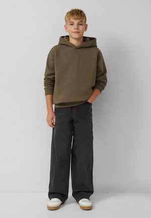 Sweatshirt à capuche vert olive, pantalon large noir, baskets blanches avec semelles beige. Design simple avec des poignets côtelés. Aucun motif visible.