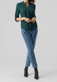Türkise Bluse mit V-Ausschnitt und hochgerollten Ärmeln, kombiniert mit hellblauen Jeans und schwarzen Stiefeletten. Glatte Textur und lockere Passform.