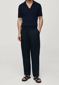Tmavě modrý pletený polo svetr s výstřihem do V, kombinovaný s volnými tmavými kalhotami a černými sandály. Hladká textura a minimalistický design.