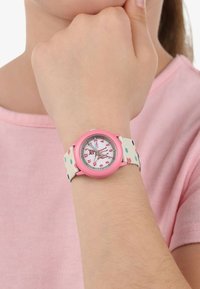 Rosa Uhr mit rundem Zifferblatt, weißen und roten Ziffern sowie einem gemusterten Stoffarmband mit bunten Punkten auf einem cremefarbenen Hintergrund.