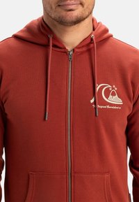 Haut à capuche rouge zippé avec une poche kangourou, des cordons de serrage et un logo représentant une vague et une montagne. Tissu doux et texturé.