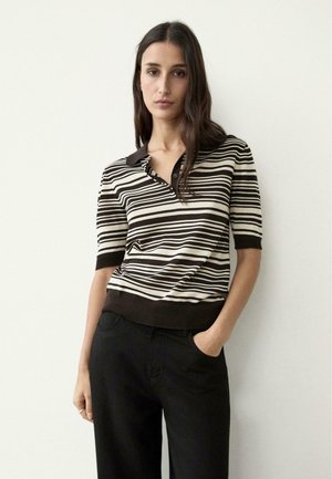 Femme aux longs cheveux noirs portant un polo à manches courtes à rayures horizontales noires et blanches et un pantalon noir, debout contre un mur blanc.