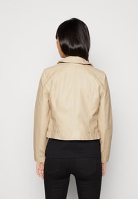 Crèmekleurig lederen jack met doorgestikte accenten op de schouders, een kraag en een cropped ontwerp. Gedragen over een zwart shirt, gezien van achteren.