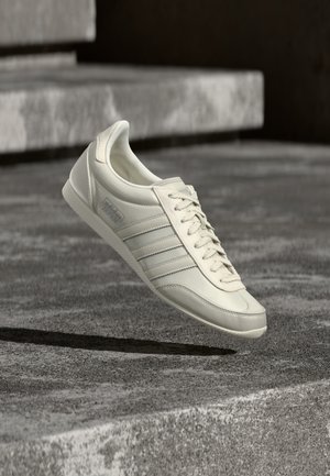 Fehér Adidas tornacipő fűzőkkel és csíkokkal, a levegőben lebegve, szürkeárnyalatos környezetben, egy texturált kősíkon és lépcsők felett.