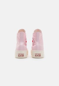 Sapatilhas de cano alto rosas feitas de tela com solas de borracha brancas. Apresentam um logo circular e "CONVERSE ALL STAR" impresso em vermelho no calcanhar.