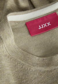 Beige Strickpullover mit Rundhalsausschnitt und einem hellgrauen Streifen am Kragen. Mit einem roten Stoffetikett mit weißem Text "JJXX."