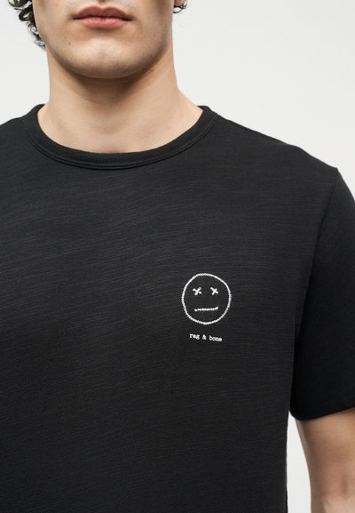 Νεαρός που φοράει μαύρο t-shirt με στρογγυλή λαιμόκοψη, με μικρό λευκό κεντημένο πρόσωπο και το κείμενο «rag & bone» στο αριστερό μέρος του στήθους.